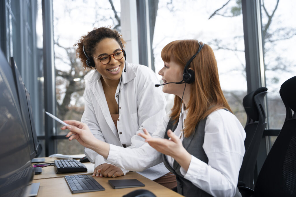 Duas mulheres trabalhando em um ciclo de atendimento. Uma delas está apontando com uma caneta para uma tela de computador enquanto mostra algo. As duas sorriem e usam fones de ouvido próprios para o atendimento ao cliente.