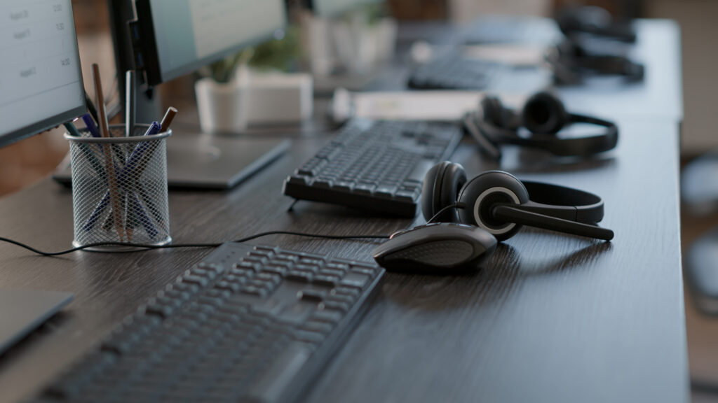 Uma grande estação de trabalho com quatro computadores, cada um com um fone headset utilizado por agentes de service desk.