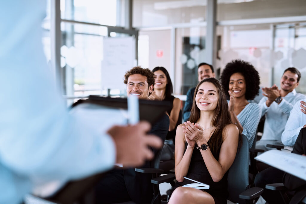 Homem usando camisa social branca, de costas para uma plateia de pessoas em treinamento. As pessoas estão em uma sala com portas e janelas de vidro, sorridentes, enquanto aplaudem o treinador.