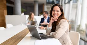Confident businesswoman with laptop on table smiling at camera while legal colleagues collaborating in the background