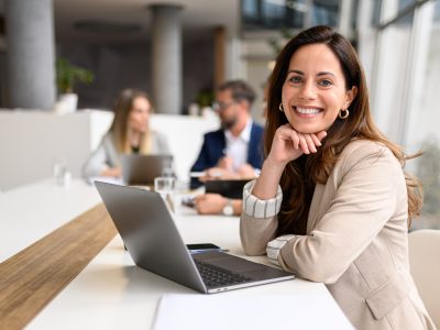 Confident businesswoman with laptop on table smiling at camera while legal colleagues collaborating in the background