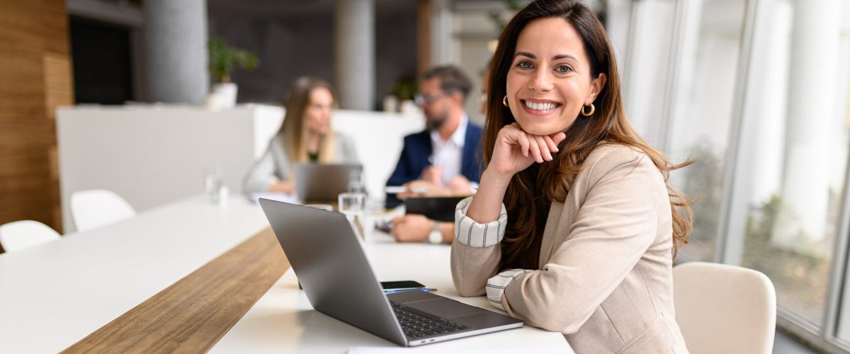 Confident businesswoman with laptop on table smiling at camera while legal colleagues collaborating in the background