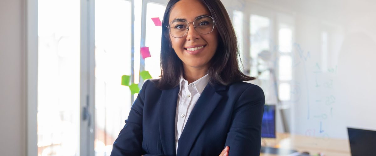 Confident Latin business leader portrait. Young businesswoman in suit and glasses posing with arms folded, looking at camera and smiling. Female leadership concept