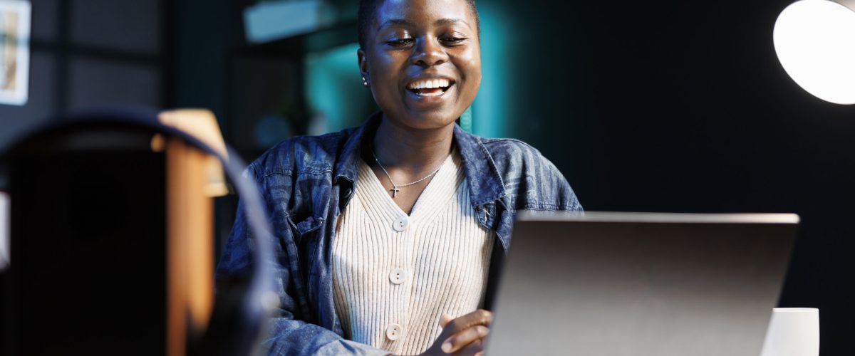 Black woman enjoying a comedy movie on her digital laptop, while seated at desk in her apartment. Excited african american lady using personal computer for virtual online communication with friends.