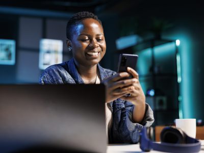 Cheerful female college student using mobile device to engage with friends on social media platforms. African american lady using cell phone to surf the web while sitting at desk in her home office.