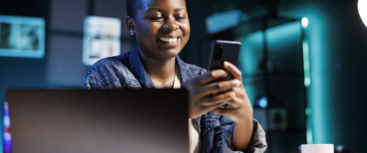 Female student using mobile device Cheerful female college student using mobile device to engage with friends on social media platforms. African american lady using cell phone to surf the web while sitting at desk in her home office.