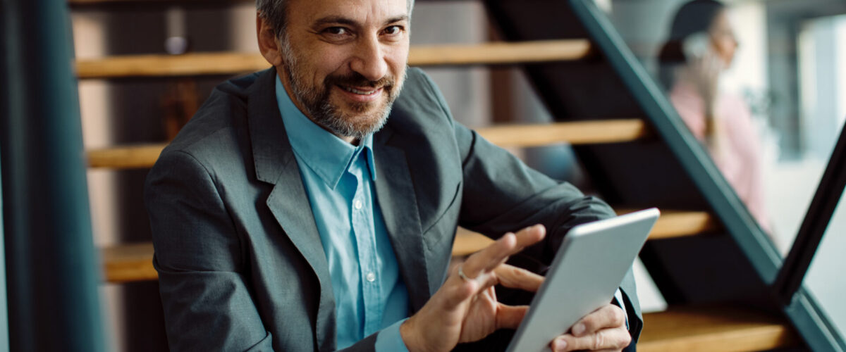 Smiling mid adult businessman using digital tablet while sitting on steps and looking at camera.