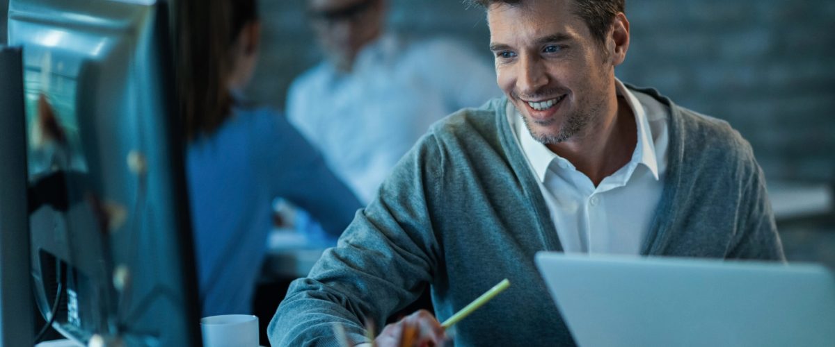 Happy male entrepreneur using desktop PC while working in the office. There are people in the background.