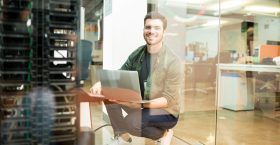 Portrait of happy young male network engineer with laptop in hand working in datacenter.