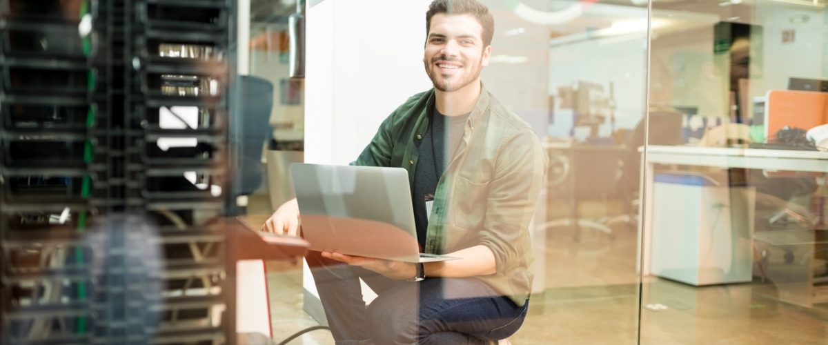 Portrait of happy young male network engineer with laptop in hand working in datacenter.