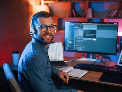 Sitting and smiling. Professional programmer is working indoors. Neon lighting.