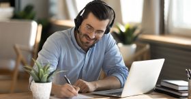 Smiling young Caucasian man in headphones glasses sit at desk work on laptop making notes. Happy millennial male in earphones watch webinar or training course or computer, study online from home.