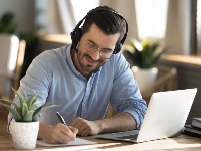 Smiling young Caucasian man in headphones glasses sit at desk work on laptop making notes. Happy millennial male in earphones watch webinar or training course or computer, study online from home.