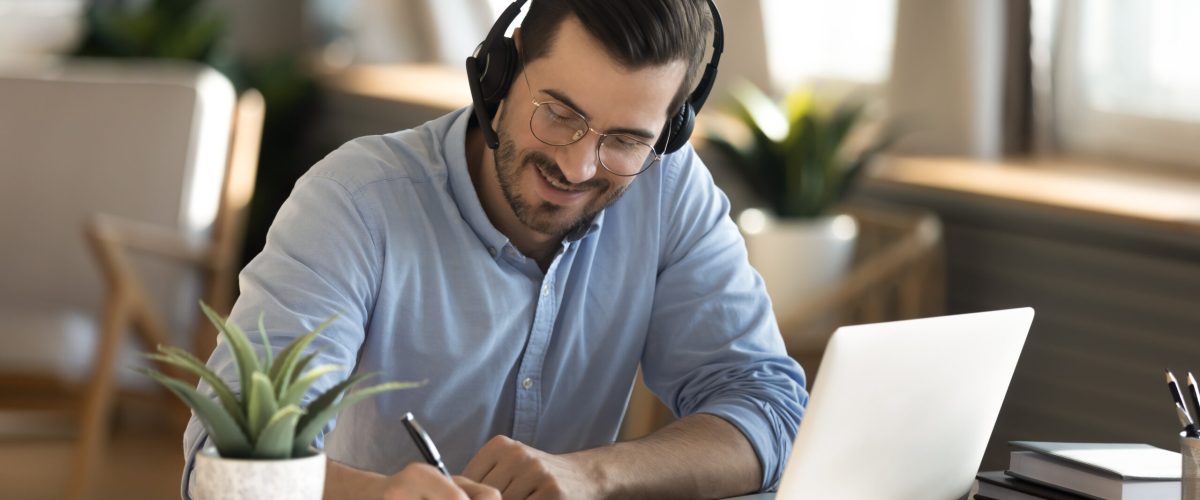 Smiling young Caucasian man in headphones glasses sit at desk work on laptop making notes. Happy millennial male in earphones watch webinar or training course or computer, study online from home.