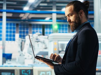 Man in photovoltaics factory monitoring system performance on laptop. Solar panels manufacturing plant employee using notebook to track energy production and efficiency, camera B close up