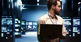 Supervisor using laptop to oversee energy consumption across server hub components and distributors. Serviceman ensuring high tech facility sensors for temperature and humidity are working properly