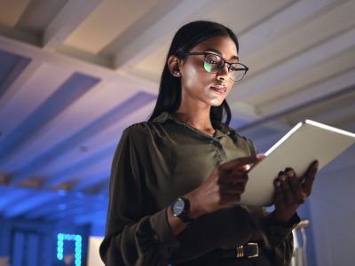 Tablet, designer and serious woman research in business startup office at night on deadline. Technology, creative professional or Indian entrepreneur reading information, email or app in neon company.